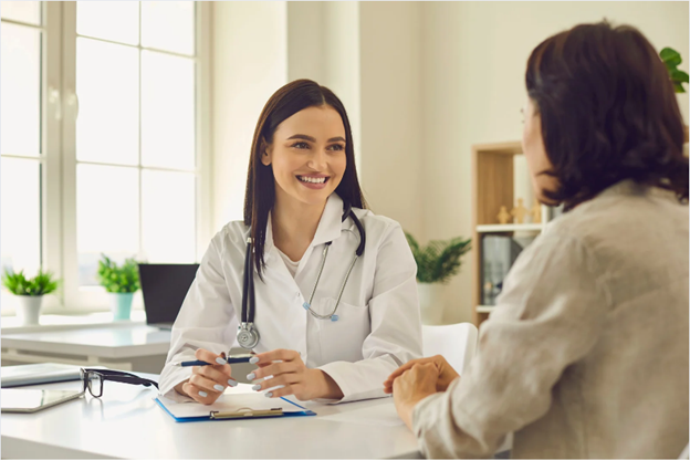 The image shows a female doctor sitting at a desk in an office, smiling and engaging with another person who appears to be a patient, while wearing a stethoscope around her neck.