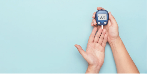 The image shows a hand holding a blood glucose monitor displaying a reading of 99, with a blue wall in the background.