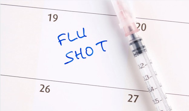 The image features a calendar page with the word  FLU SHOT  written on it, accompanied by a syringe and a pen.