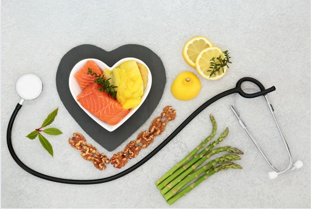 The image features a heart-shaped plate with various foods, including fish, lemon wedges, and vegetables, surrounded by medical items such as a stethoscope, pills, and a blood pressure cuff, all arranged on a surface that resembles a table with a white tablecloth.