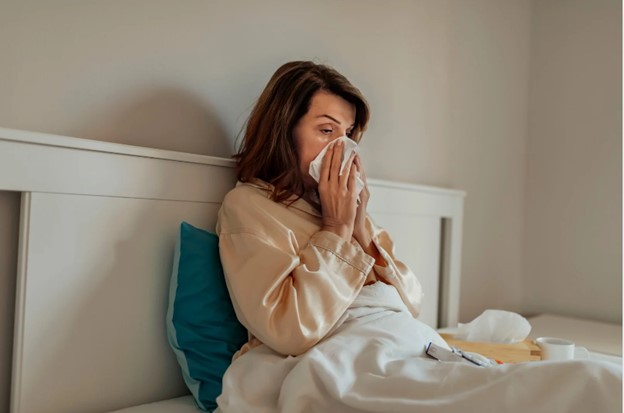 A woman lies in bed with her head covered by a pillow, holding a tissue to her nose, appearing to be crying or sneezing.