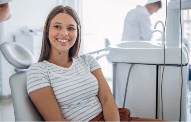 The image shows a young woman sitting in a dentist s chair with a smile on her face, surrounded by dental equipment.