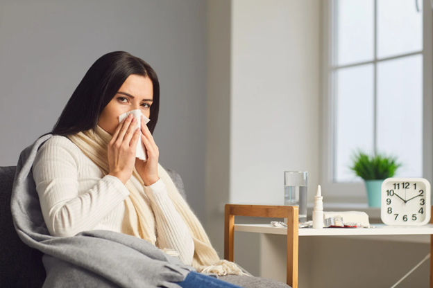 The image shows two photos side by side  one of a woman with her eyes closed while holding a tissue to her nose, and another of a person seated at a desk with a cup and a clock, also holding a tissue to their nose. Both are indoors with a blurred background that suggests a domestic setting.