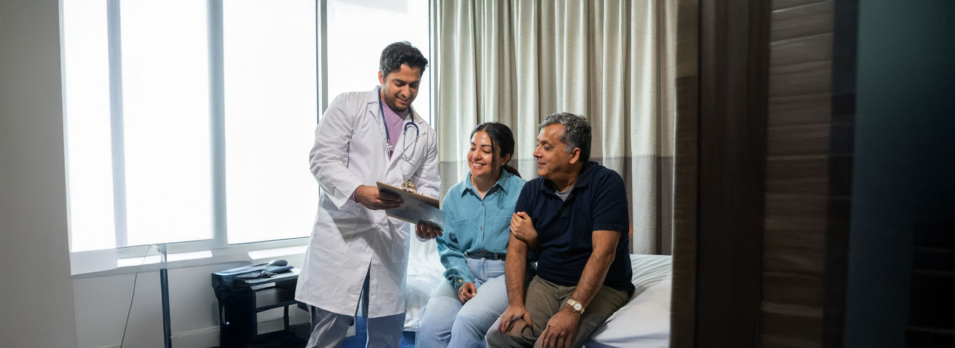 The image shows three individuals in a room with medical equipment one person appears to be a healthcare professional holding a tablet, another seems to be a patient, and a third might be a visitor or family member. They are all engaged in an interaction that could involve sharing information from the tablet.