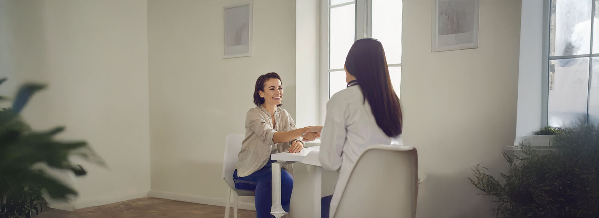 In the image, two individuals are engaged in conversation at a table within a room with white walls and a window, while another person sits nearby observing their interaction.