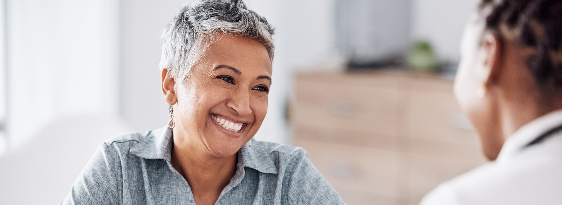 The image depicts a woman smiling at a person seated across from her, both are indoors with a blurred background suggesting an office setting.