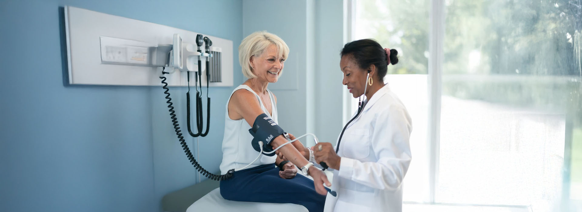 A woman sits on a medical examination table with a blood pressure cuff around her arm, receiving attention from a standing healthcare professional wearing a stethoscope.