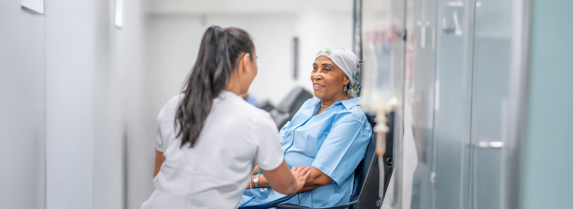 The image shows two individuals engaged in conversation within a hospital setting, with one person seated on a chair and another standing, both wearing medical scrubs.
