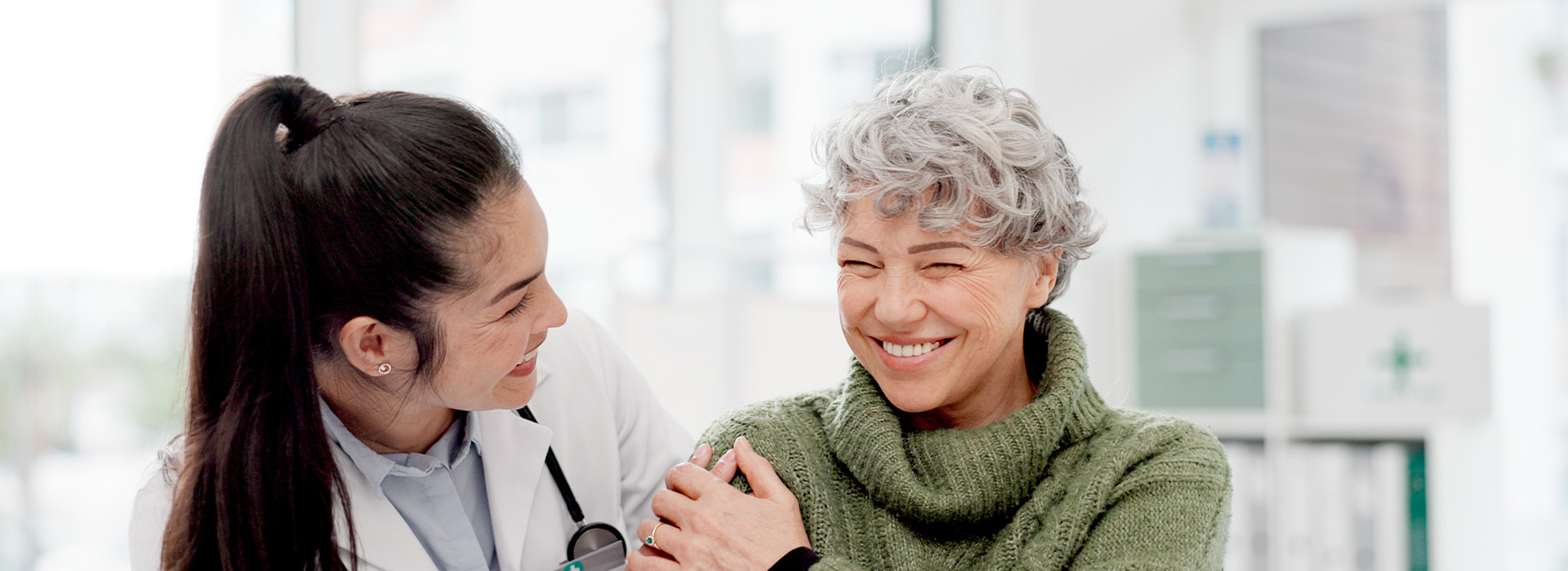 Two individuals are standing close together, smiling at each other. The person on the left appears to be a woman wearing a white lab coat, while the individual on the right seems to be an older adult. They are both indoors, and there is a blurred background suggesting they might be in a public building with some greenery visible.