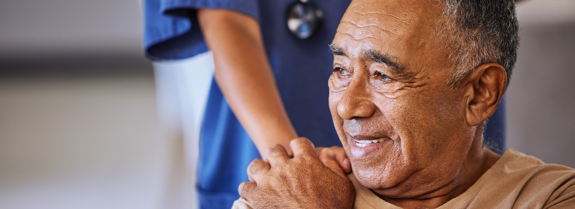 The image shows an elderly man with a smile on his face, wearing glasses and seated in a wheelchair, being assisted by someone standing beside him who appears to be a healthcare professional.