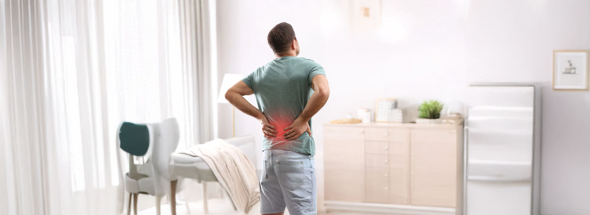 The image depicts a man standing in a kitchen with his back turned to the camera, wearing a green shirt, and looking out towards a living room area where he appears to be experiencing discomfort or pain in his lower back.