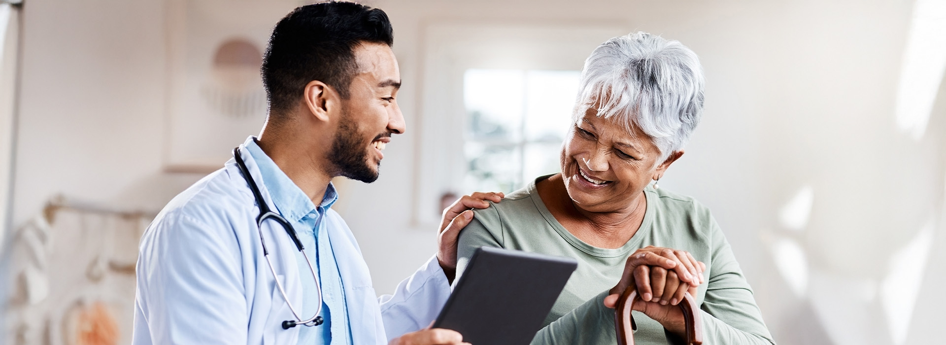 The image shows two individuals in a medical setting, with one person holding a tablet and smiling at an elderly patient who appears to be receiving care or advice.