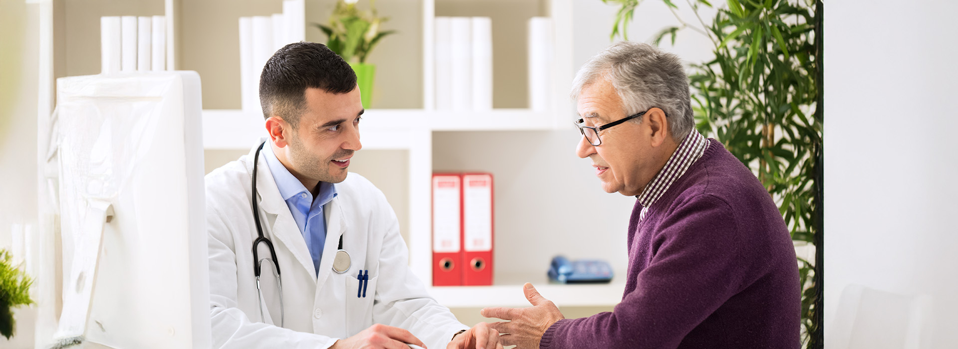 The image shows two elderly individuals in a medical setting, with one person wearing a white coat and stethoscope, likely a healthcare professional, engaging in conversation with another individual who appears to be receiving care or advice.