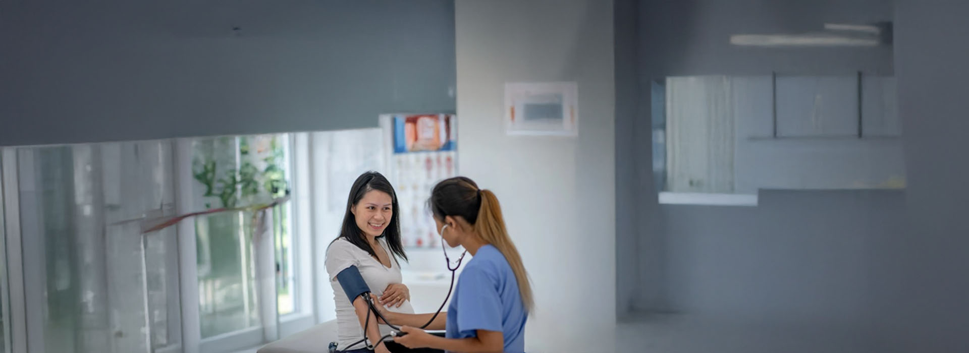 A woman wearing a white shirt stands next to a man in scrubs who is holding a clipboard with papers, both inside a hospital room.