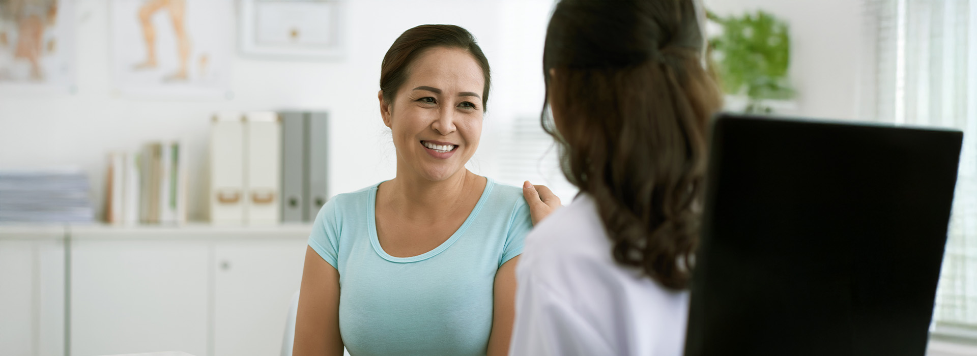 The image shows a woman sitting at a desk with a laptop, engaged in conversation with another person who appears to be standing behind her.