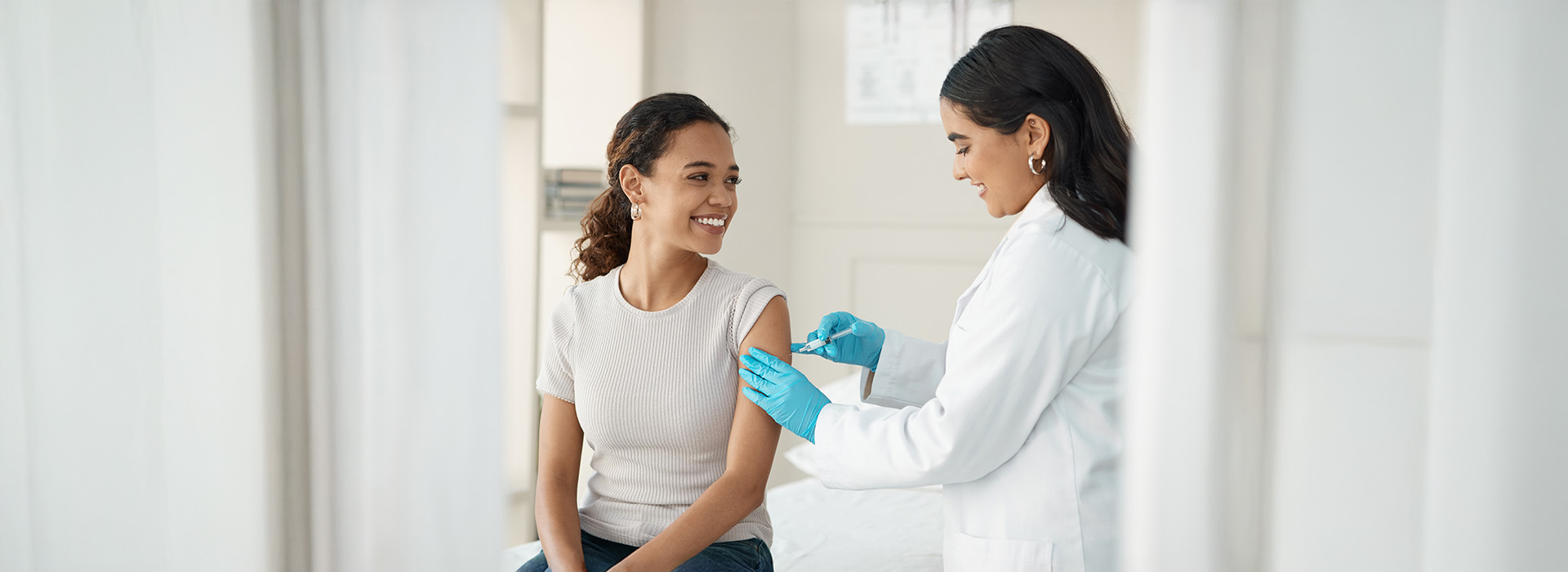 The image shows a woman receiving a COVID-19 vaccination from a healthcare professional in a medical setting.