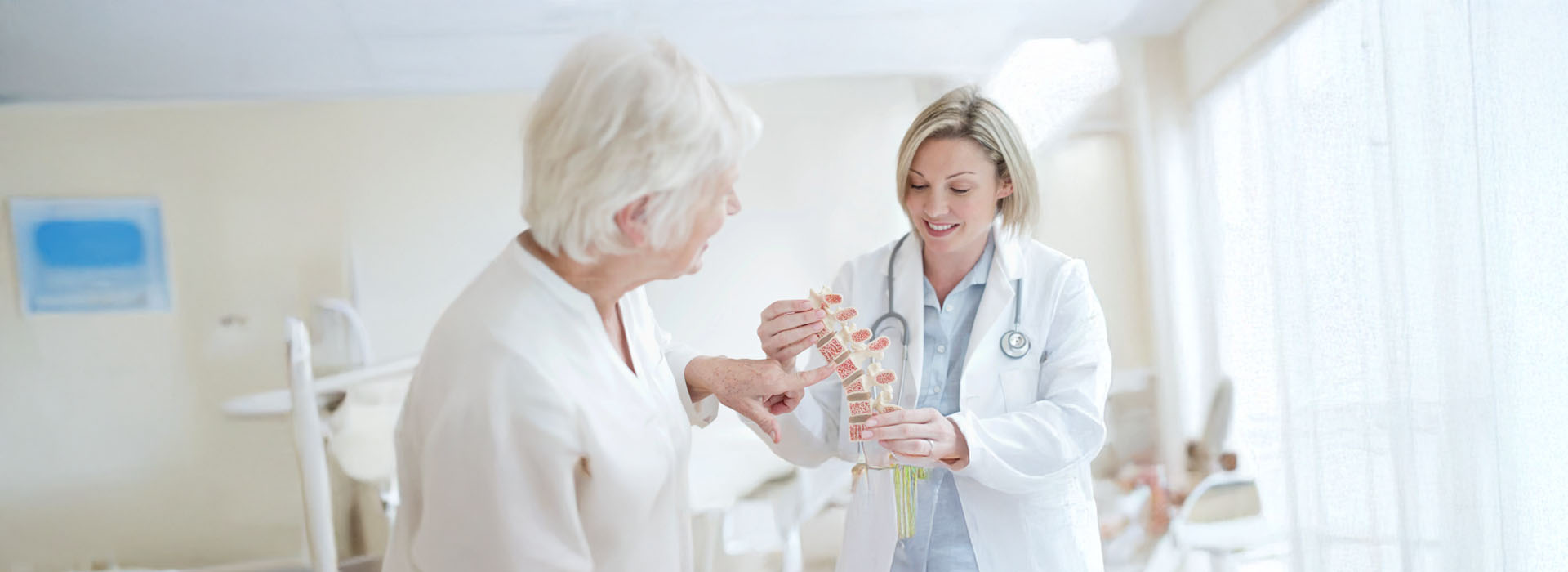 The image shows two older adults with white hair receiving medical attention from a female healthcare professional wearing a blue scrub top in a hospital setting.
