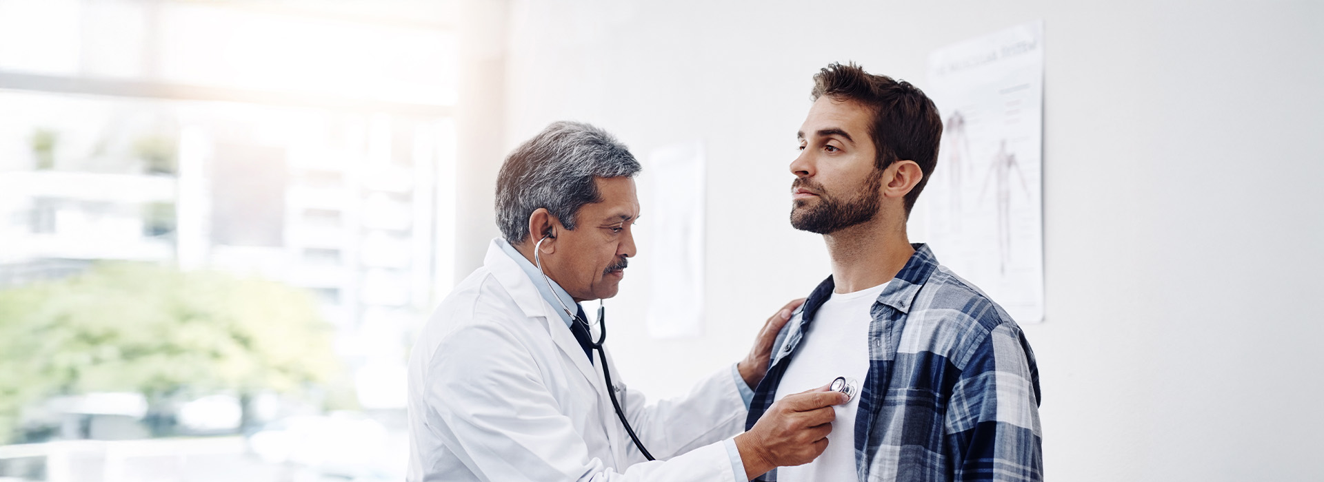 A man with a stethoscope around his neck is examining another man who is seated, while a third man stands nearby observing. The setting appears to be a medical office.