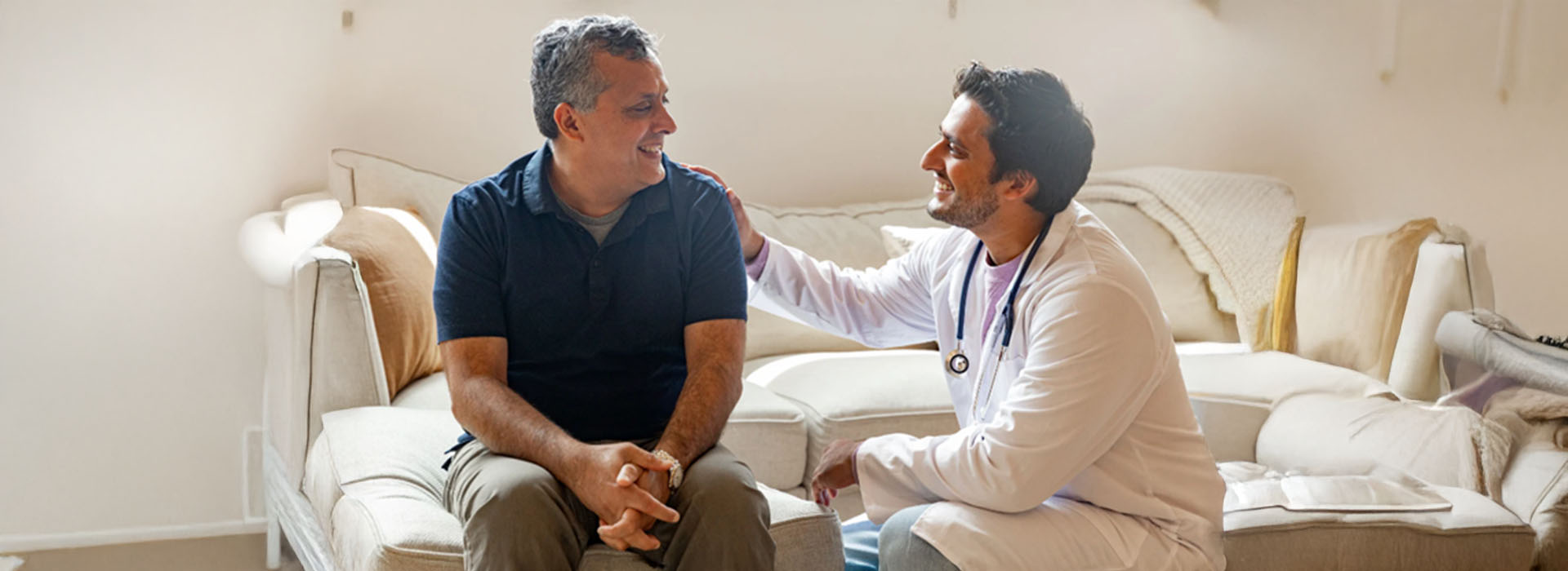 A man and a woman are sitting on a couch, with a third person standing between them, engaging in conversation. The setting appears to be indoors, possibly a living room, with a couch and a television visible in the background.