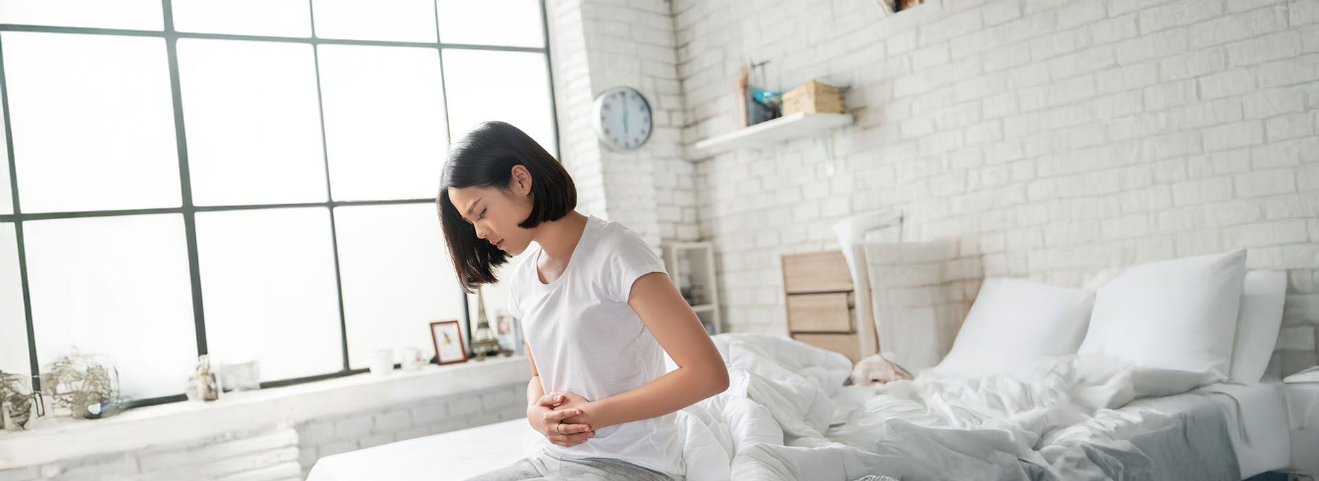 A woman stands in a modern bedroom with a neatly made bed, looking down at her hands clasped together.