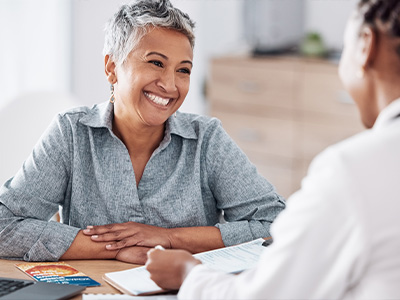 The image shows a woman sitting at a desk with a laptop, smiling and looking towards a person who appears to be speaking with her, while another individual is seated across from them, engaged in conversation.