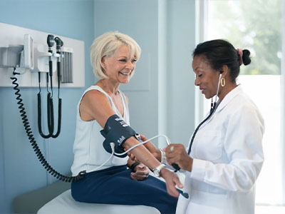 A woman sits on a hospital bed with an IV drip while another woman, possibly a nurse, checks her blood pressure using a stethoscope.