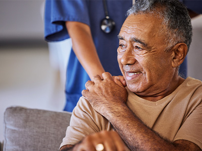 The image shows a man sitting on a couch with his hands clasped together, wearing a white shirt and smiling at the camera. He appears to be an elderly individual with a mustache and is surrounded by people who seem to be engaged in conversation or activity around him.