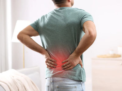 An adult male with back pain, standing in a room, leaning forward slightly, wearing a green t-shirt, blue jeans, and a red band around his waist.