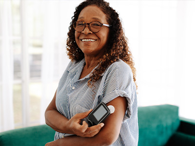 A smiling older woman holding a cell phone with her left hand while standing in a room with a couch visible in the background.
