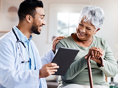 The image depicts two individuals engaged in an interaction, with one person holding a tablet and smiling at the camera while another person, who appears to be elderly, is being assisted by a healthcare professional wearing a stethoscope, both within a domestic setting.
