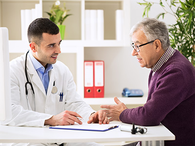 A man in a white coat, standing at a desk with a clipboard, is engaged in conversation with an older gentleman who appears to be a patient.