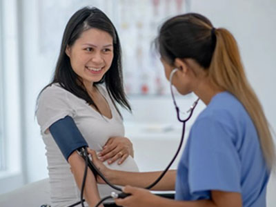 A woman wearing a white shirt is receiving a blood pressure check from a medical professional using a stethoscope while seated in a hospital setting.