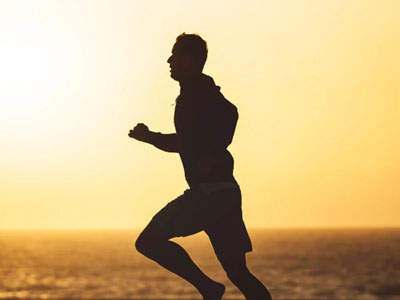 The image shows a person running on a beach at sunset with the silhouette of their body against the bright sky, captured from behind.