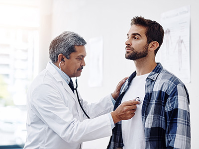 The image shows a man wearing a plaid shirt receiving a medical examination from a doctor in a professional setting.