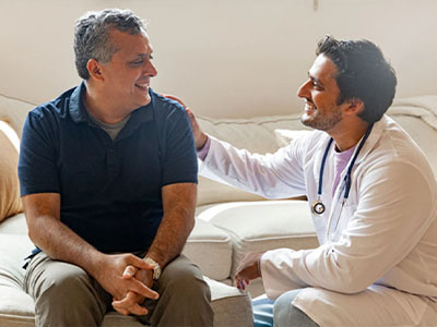 The image shows two men sitting on a couch  one appears to be a medical professional wearing a white coat with a stethoscope around his neck, smiling at the other man who is seated next to him.