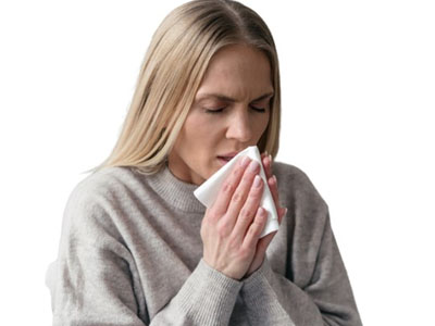 The image shows a woman with blonde hair wearing a gray sweater, holding her face with both hands, and appears to be sneezing or experiencing discomfort.