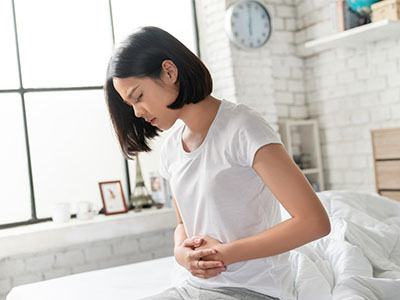A person with dark hair wearing a white shirt is sitting up with their hand on their chest, appearing to be experiencing discomfort or pain.