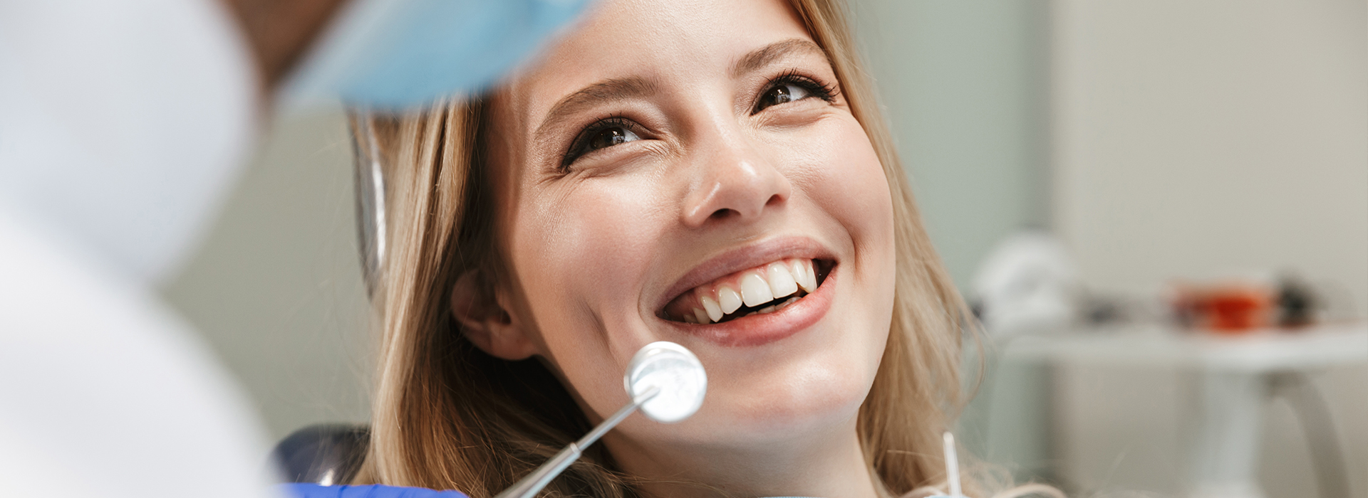 A woman with a bright smile sits in a dental chair, receiving dental care from a professional wearing a blue surgical mask.
