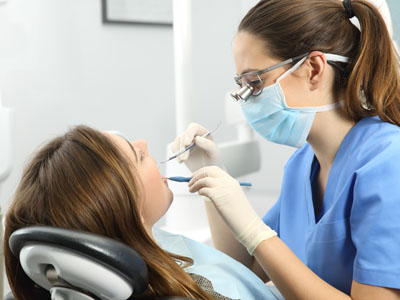 A dental hygienist is performing an oral procedure on a patient, with both individuals wearing face masks and the hygienist using specialized tools.