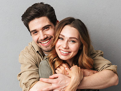 A man and woman embrace each other warmly, both smiling broadly, against a neutral background.