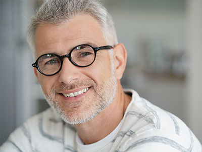 The image features two side-by-side photos of a man with glasses, smiling and looking directly at the camera. He has short hair and a beard, and he appears to be indoors, possibly in a room with a white wall in the background.